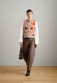 Többszínű mellény geometriai mintákkal fehér hosszú ujjú ing felett, barna nadrággal és fekete bokacsizmával párosítva. Barna válltáska.