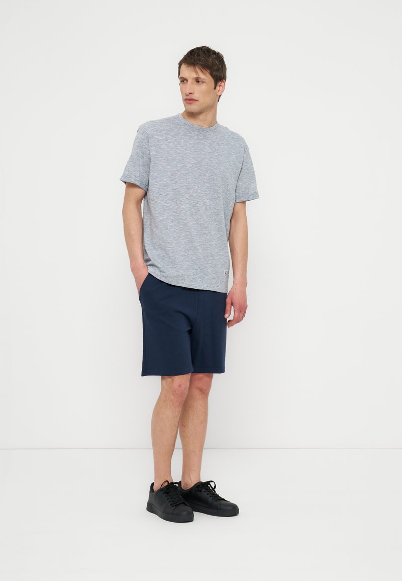 Homme debout les mains dans les poches portant un t-shirt gris clair à manches courtes, un short bleu marine et des baskets noires sur fond blanc.