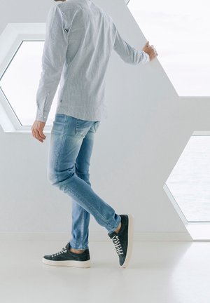 Man in gestreept overhemd en spijkerbroek leunt op muur naast zeshoekige ramen, licht een voet iets op, binnen met natuurlijk licht.