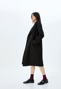 Abrigo largo negro de diseño formal, hecho de un material similar a la lana. Presenta un cierre de un solo botón y solapas anchas. Se lleva sobre una blusa clara, acompañado de zapatos negros y calcetines burdeos.