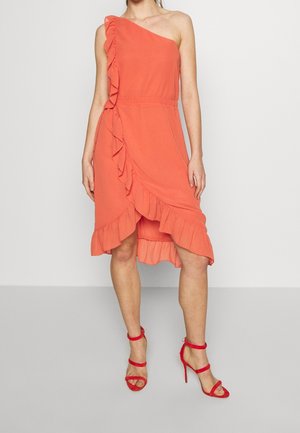 Robe corail sans manches avec une épaule, détails volants, ourlet asymétrique, portée avec des sandales rouges à talons hauts et bride à la cheville.