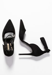 Černé semišové boty na vysokém podpatku s špičatými špičkami, vykrojenými stranami a kotníkovým páskem. Součástí jsou lesklý tenký podpatek a hladký kožený interiér.