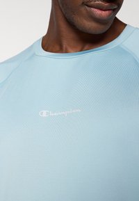 Ανοιχτό μπλε αθλητικό πουκάμισο από ομαλό ύφασμα, με στρογγυλή λαιμόκοψη και λευκό λογότυπο "Champion" στην αριστερή πλευρά του στήθους.