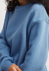 Persona con un sudadera azul de corte holgado, con puños y cuello acanalados, brazos cruzados, frente a un fondo claro y liso.