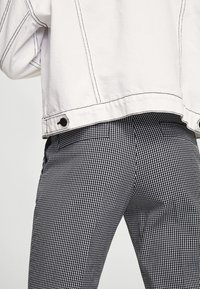 Veste en denim blanche avec détails de couture noire sur un pantalon à carreaux noir et blanc. Le pantalon a une coupe ajustée et des poches arrière.