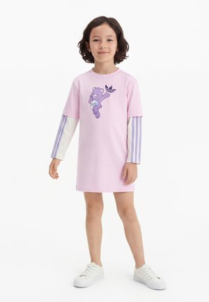 Copil în picioare, purtând o rochie roz cu un desen cu urs violet și mâneci lungi suprapuse cu dungi violet, asortată cu adidași albi.