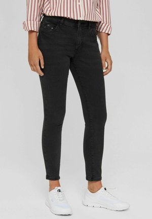 Persona che indossa jeans neri aderenti, sneakers bianche e una camicia a maniche lunghe a righe arrotolata, in piedi davanti a uno sfondo semplice.