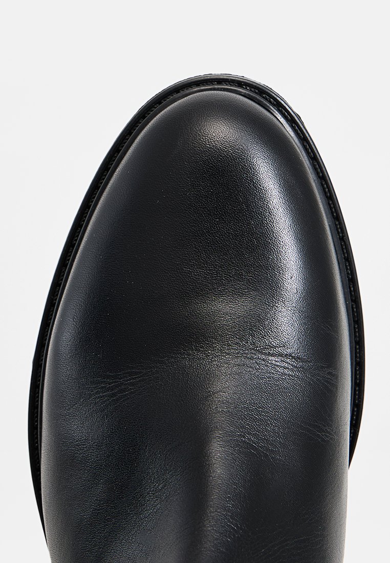 Zapato de cuero negro con punta redondeada, textura suave y una fina costura decorativa a lo largo del borde de la suela.