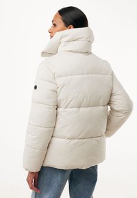 Donna che indossa un piumino beige chiaro con colletto alto e jeans blu, vista di spalle e con lo sguardo rivolto a sinistra.