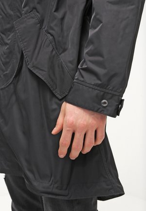 Gros plan d’une personne portant un manteau noir, montrant un poignet de manche avec un bouton-pression en métal et une main reposant le long du corps.