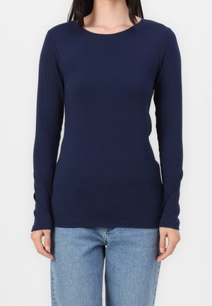 Femme portant une chemise bleu marine ajustée à manches longues et un jean bleu clair, debout devant un fond blanc uni.