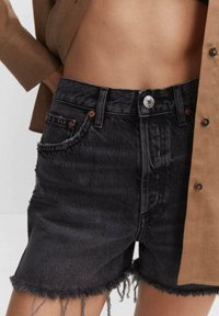 Pantalones cortos de mezclilla negra con dobladillo deshilachado, diseño de cinco bolsillos, cierre de botón plateado y detalles de costura en contraste. Acentuado con una blusa marrón clara.