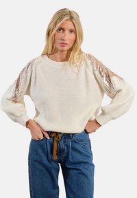 Maglione lavorato a maglia color crema con dettagli in paillettes sulle maniche, scollo rotondo e vestibilità rilassata. Abbinato a jeans blu a vita alta.