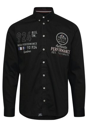 Camisa negra de manga larga con cuello abotonado, costuras decorativas y varios logotipos bordados en el pecho y en la manga.