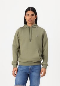 Hoodie en coton vert olive avec une poche frontale, un capuchon ajustable avec cordon de serrage, et un petit logo brodé sur la poitrine. Coupe décontractée.