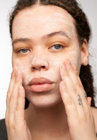 Une personne appliquant un nettoyant pour le visage, avec une texture mousseuse sur sa peau, affichant un teint clair, des tatouages et des boucles légères dans un cadre neutre.