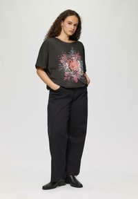 Schwarzes T-Shirt mit floraler Gestaltung in Rosa und Weiß, kombiniert mit lockeren schwarzen Hosen und schwarzen Stiefeletten. Modell steht mit den Händen in den Taschen.
