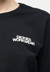 Μαύρο πουκάμισο με στρογγυλή λαιμόκοψη και λευκό λογότυπο "Dickies Workwear" τυπωμένο στο πάνω αριστερό μέρος του στήθους, το οποίο φοράει ένα άτομο.
