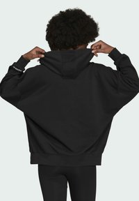 Felpa nera oversize con un grande cappuccio, polsini a coste e un discreto dettaglio del logo sulla manica. Il tessuto sembra morbido e strutturato.