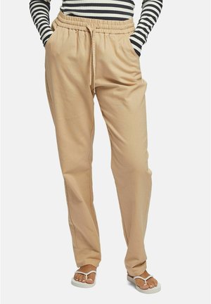 Pantalons beiges légers, à cordon de serrage et à coupe décontractée. Dotés d'une taille élastique et de poches latérales. Associés à des sandales blanches.
