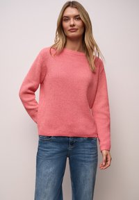 Pull en maille rose clair à col arrondi et manches longues, associé à un jean denim bleu, sur un fond neutre.