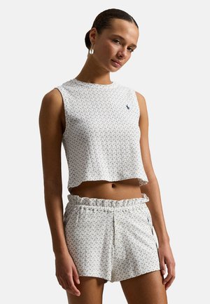 Femme portant un haut blanc sans manches court et un short assorti à taille volantée avec un motif géométrique subtil, coiffée de boucles d'oreilles en perles.