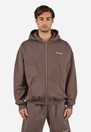 Rjava zip-up kapuca z dvema sprednjima žepoma, rebrastimi manšetami in logotipom na prsih. Narejena iz mehkega, debelega materiala z udobnim, sproščenim krojem.