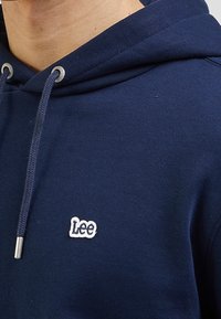 Felpa blu navy realizzata in morbido tessuto. Presenta un cordino argentato e un piccolo logo bianco "Lee" sul davanti. Texture liscia.