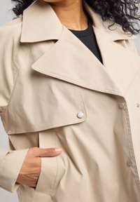 Beige trench coat dengan kerah lebar, desain double-breasted, dan saku samping. Tekstur halus dengan kancing tekan perak.