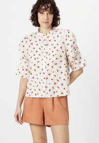 Floral blouse in cream with short sleeves, featuring red rose patterns, button front closure, and slight texture; paired with orange shorts.