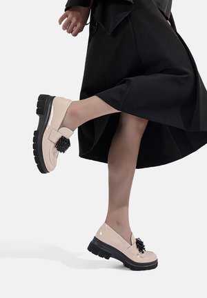 Scarpe in pelle lucida beige con suola nera spessa, con un pompon nero decorativo sulla parte superiore. Indossate con una gonna nera fluida.