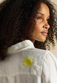 Valge kraega särk, millel on kollase päikese tikand seljal. Kangas on kerge ja veidi tekstureeritud viimistlusega.