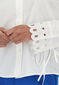 Camicia bianca con bottoni, caratterizzata da dettagli a taglio intricati sulle maniche a scollo scallop e lacci in vita, realizzata in tessuto leggero.