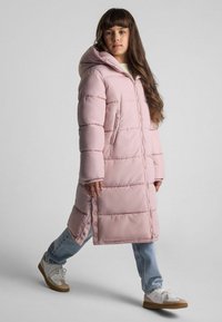 Lång rosa vadderad kappa med huva, med sydda paneler, dragkedjestängning och sidoknappar, matchad med denim och avslappnade sneakers.