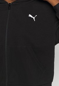 Sweat à capuche noir zippé avec logo Puma blanc sur la poitrine et poche kangourou à l'avant, porté par une personne ajustant la capuche sur le côté droit.