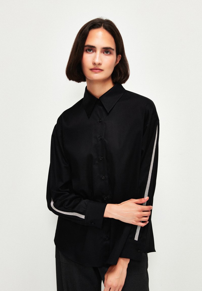 Camicia nera con colletto, caratterizzata da un design elegante e dettagli argentati lungo le maniche. Realizzata in tessuto morbido.