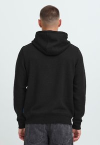 Uomo con capelli corti che indossa un hoodie nero e jeans grigio scuro, visto di spalle su uno sfondo leggero e uniforme.