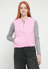 Rosa fleecevest med teksturert finish, med frontlukking og to sidelommer, brukt over en grå langermet skjorte og mørke bukser.