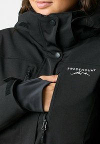 Zwarte buitenjas met een hoge kraag, rits- en klittenbandzakken, voorzien van een logo. De stof lijkt glad met een matte textuur.