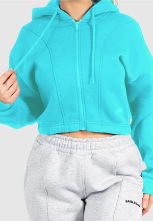Personne portant un sweat à capuche court turquoise vif et un pantalon de survêtement gris clair à taille élastique avec un petit logo noir sur la cuisse gauche.