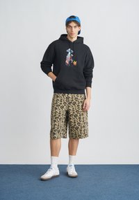 Μαύρο φούτερ με κουκούλα, εκτύπωση και τσέπη τύπου καγκουρό, σε συνδυασμό με μπεζ σορτς με λεοπάρ σχέδιο. Λευκά αθλητικά παπούτσια με γκρι λεπτομέρειες και καλτσάκια.