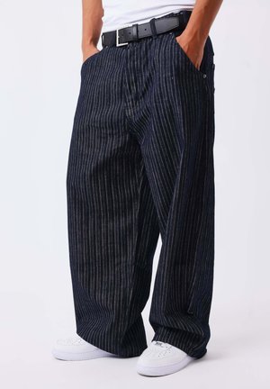 Personne portant un pantalon large à fines rayures foncées, une ceinture noire, des baskets blanches et une chemise blanche, les mains dans les poches, sur un fond uni.