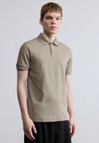 Polo bege de manga curta, em algodão, com gola clássica e dois botões. Logo discreto no peito esquerdo, textura suave.