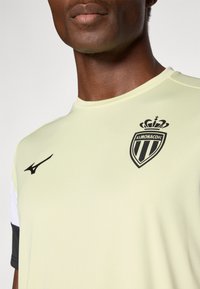 Svetlo rumena športna majica s kratkimi črnimi in belimi rokavi, ki na prsih vsebuje črni logotip AS Monaco in logotip Mizuno.