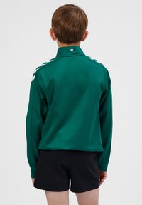 Grøn langærmet skjorte med høj krave, med hvide accentstriber på skuldrene, lavet af glat stof. Båret med sorte shorts.