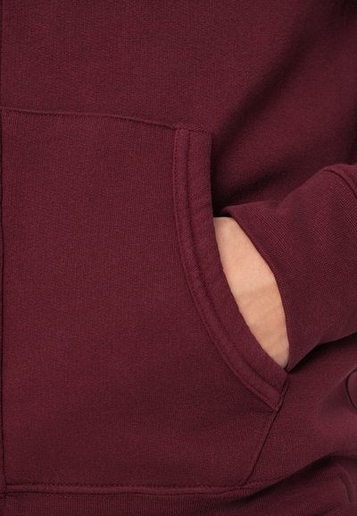 Ruka djelomično unutar prednjeg džepa maroon dukserice koja prikazuje teksturu tkanine i detalje šivanja.