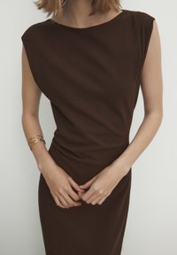 Robe ajustée marron avec un col rond et des détails drapés, fabriquée dans un tissu lisse. Les accessoires comprennent plusieurs bracelets en or sur un poignet.