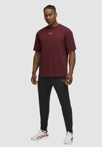 Bordeauxfarbenes T-Shirt mit kurzen Ärmeln und weißem Logo, kombiniert mit schwarzen Jogginghosen. Das Schuhwerk hat weiße und pastellfarbene Akzente.