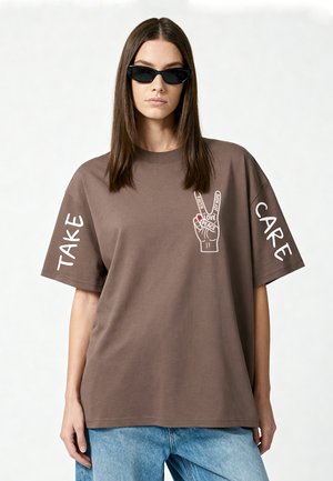 Donna con capelli castani lisci e occhiali da sole neri che indossa una maglietta marrone oversize con "TAKE CARE" sulle maniche e una grafica di una mano della pace.