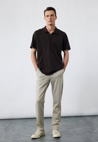 Mann steht mit Händen in den Taschen, trägt ein schwarzes kurzärmliges Poloshirt, beige Hosen und beigen Sneakers vor einem schlichten Hintergrund.